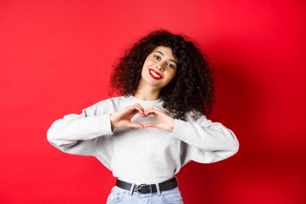 Belle jeune femme aux cheveux bouclés montrant le geste du cœur, dire je t'aime et sourire romantique à la caméra, debout sur fond rouge.