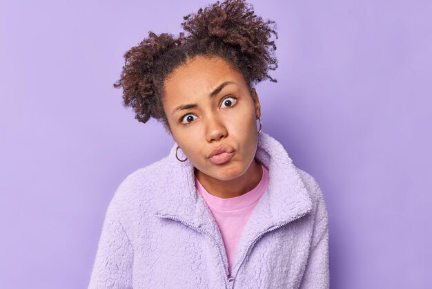 Belle jeune femme aux cheveux bouclés regarde avec un regard attentif la caméra moue les lèvres concentrées sur quelque chose vêtu d'une veste de fourrure chaude écoute attentivement les informations isolées sur un mur violet.