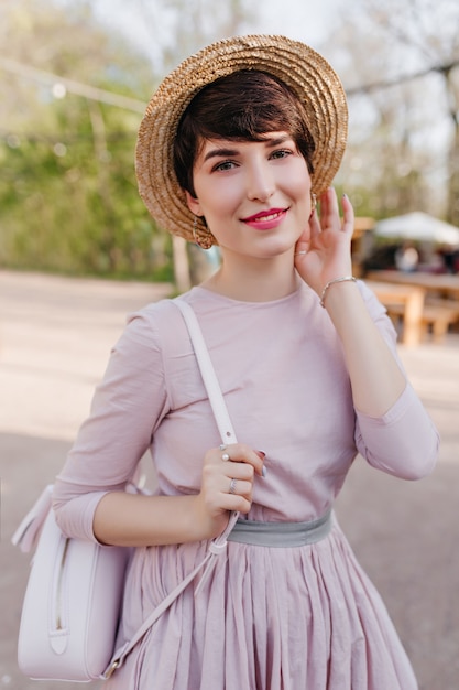 Belle jeune femme aux cheveux courts brillants posant avec plaisir pendant la promenade dans le parc