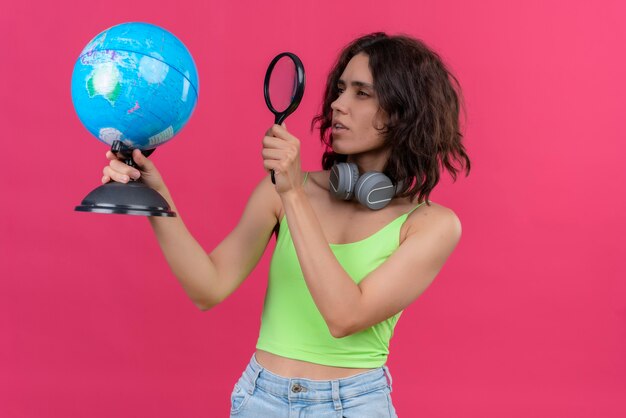 Une belle jeune femme aux cheveux courts en vert crop top dans les écouteurs regardant attentivement globe avec loupe