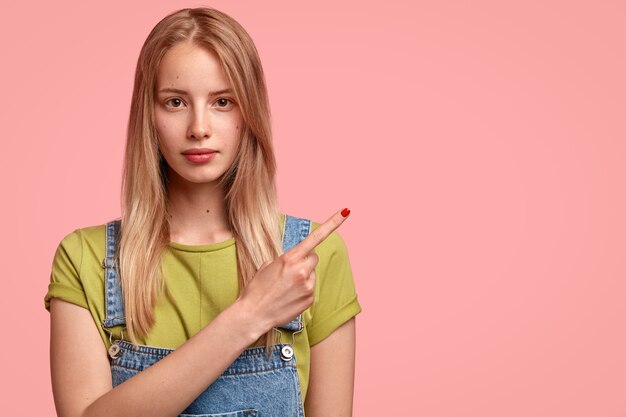 Belle jeune femme aux cheveux longs, pointe de côté avec l'index dans le coin supérieur droit, vêtue d'une salopette en denim