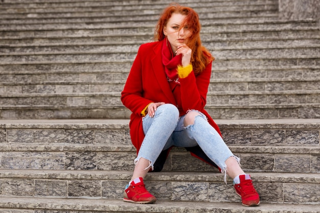 Photo gratuite une belle jeune femme aux cheveux rouges dans un manteau rouge et un jean est assis sur les marches à l'extérieur