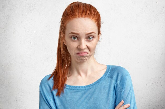 Belle jeune femme aux cheveux rouges mécontente de quelque chose, courbe les lèvres et a une expression de mécontentement, exprime la négativité