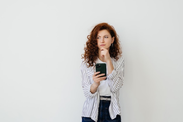 Belle jeune femme aux longs cheveux roux naturels pensant à l'expression du visage et à la pose tenant un téléphone portable