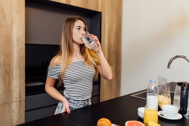 Belle jeune femme blonde buvant de l'eau dans la cuisine avec des aliments sains sur la table