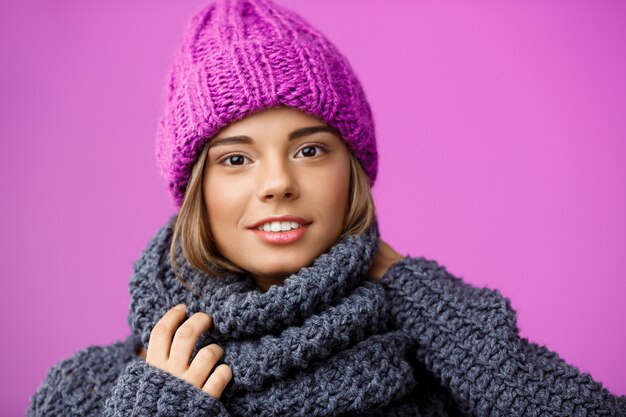 Belle jeune femme blonde en chapeau et pull en tricot souriant sur violet.