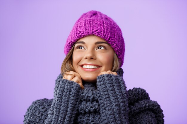 Belle jeune femme blonde en chapeau et pull tricoté souriant à côté sur violet.