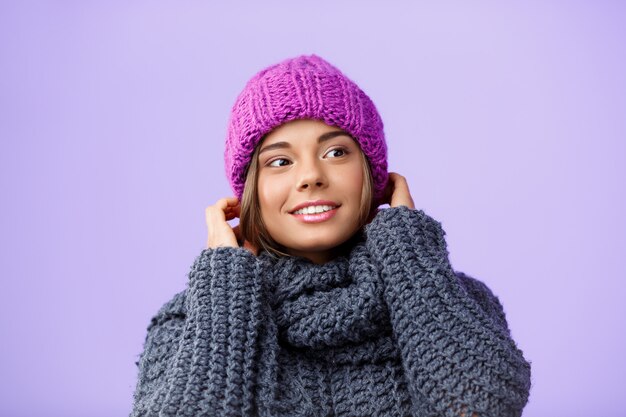 Belle jeune femme blonde en chapeau et pull tricoté souriant à côté sur violet.