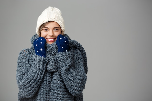 Belle jeune femme blonde en pull bonnet et mitaines souriant sur gris.