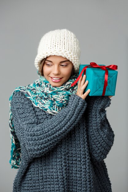 Belle jeune femme blonde en pull bonnet tricoté et écharpe souriant tenant boîte cadeau sur fond gris.