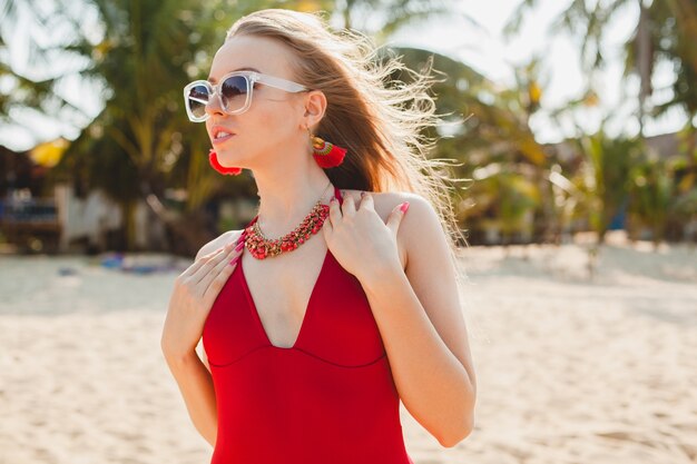 Belle jeune femme blonde se faire bronzer sur la plage en maillot de bain rouge, lunettes de soleil