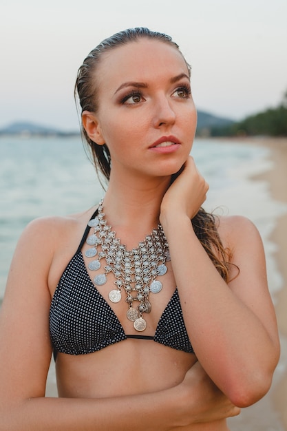 Belle jeune femme blonde se faire bronzer sur la plage de sable en maillot de bain bikini, collier vintage