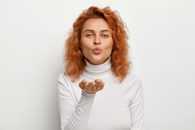 Photo gratuite belle jeune femme bouclée rousse souffle air baiser à la caméra, fait mwah, garde les lèvres pliées