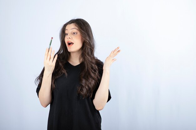 Belle jeune femme avec une brosse cosmétique debout sur un mur blanc.