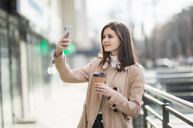 Belle jeune femme brune avec café à emporter prenant selfie