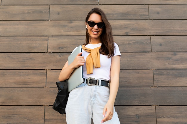 Belle jeune femme brune charmante et charmante souriante regardant la caméra tenant un ordinateur portable et des lunettes de soleil noires en t-shirt blanc et un jean bleu clair dans la rue près du mur marron.