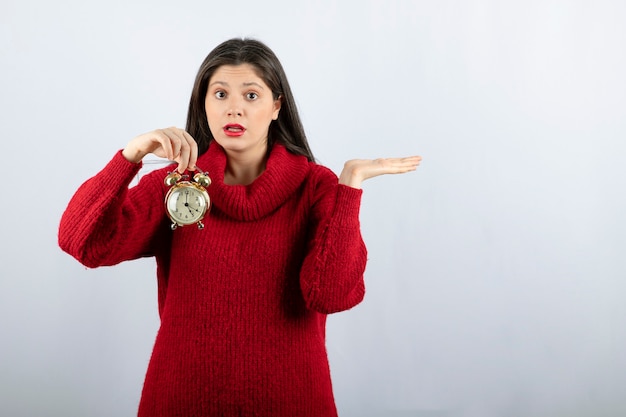 Belle jeune femme brune tenant un réveil debout sur fond blanc