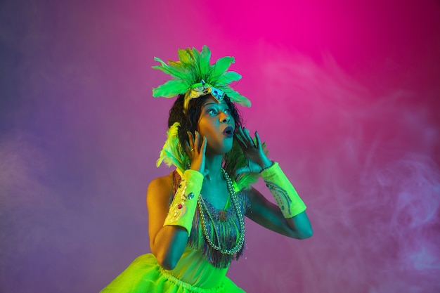 Photo gratuite belle jeune femme en carnaval, costume de mascarade élégant avec des plumes dansant sur un mur dégradé en néon