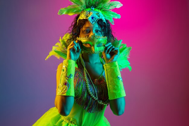 Belle jeune femme en carnaval, costume de mascarade élégant avec des plumes dansant sur un mur dégradé en néon