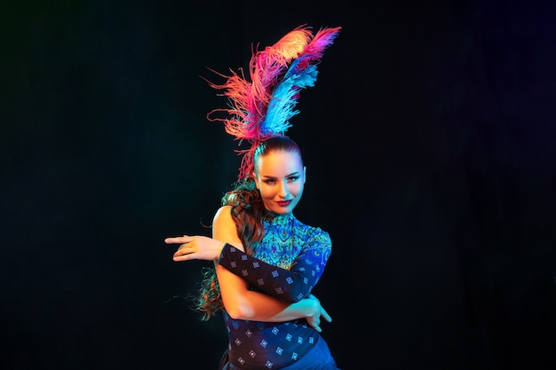 Belle jeune femme en carnaval, élégant costume de mascarade avec des plumes sur un mur noir en néon