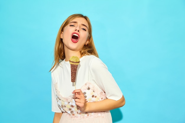 Belle jeune femme chantant avec des accessoires faux microphone. Femme à la mode dans des vêtements d'été décontractés. Modèle drôle isolé sur mur bleu