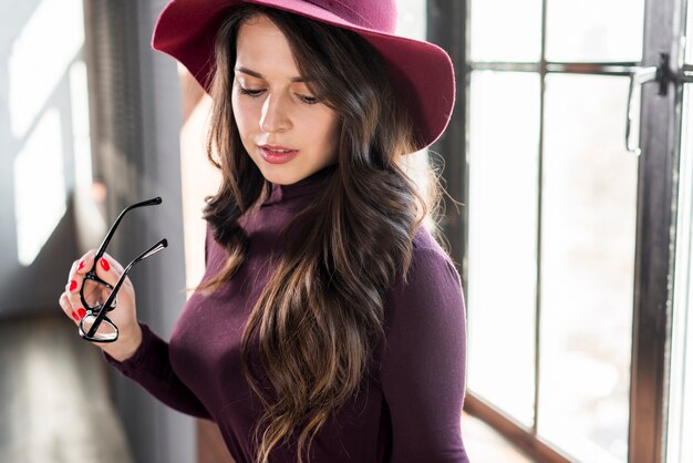 Une belle jeune femme avec un chapeau rose tenant des lunettes à la main, regardant vers le bas