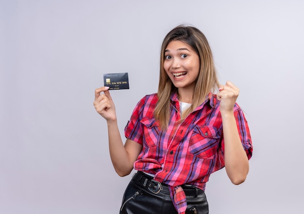 Une belle jeune femme en chemise à carreaux souriant tout en montrant la carte de crédit avec le poing fermé sur un mur blanc
