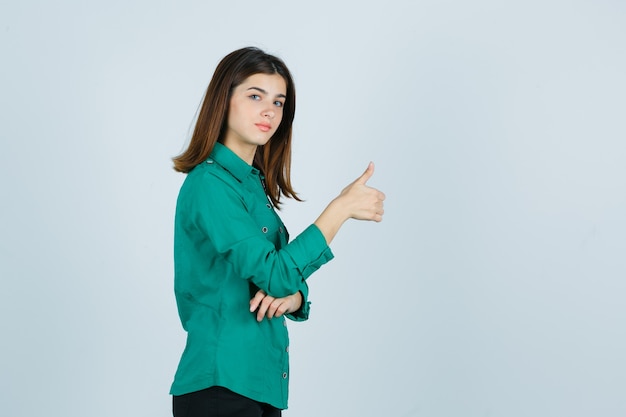 Belle jeune femme en chemise verte montrant le pouce vers le haut et à la recherche de confiance.