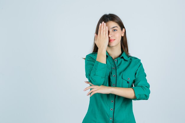 Belle jeune femme en chemise verte tenant la main sur les yeux et à l'optimiste, vue de face.