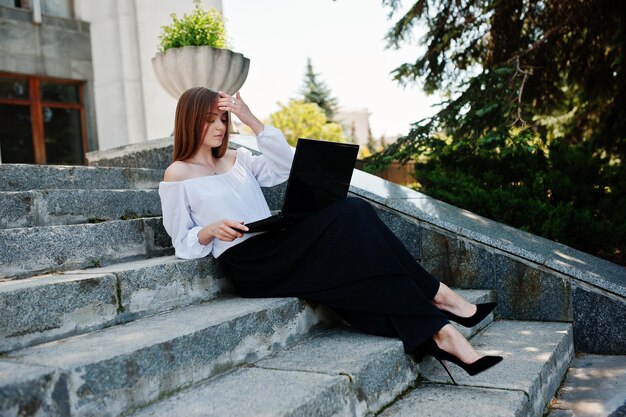 Belle jeune femme en chemisier blanc large pantalon noir et talons hauts classiques noirs assis dans les escaliers et travaillant sur son ordinateur portable