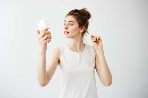 Belle jeune femme avec chignon en regardant l'écran du téléphone corrigeant les cheveux sur fond blanc.
