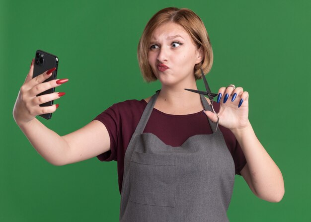 Belle jeune femme coiffeur en tablier à l'aide de smartphone faisant selfie avec des ciseaux étant confus debout sur le mur vert