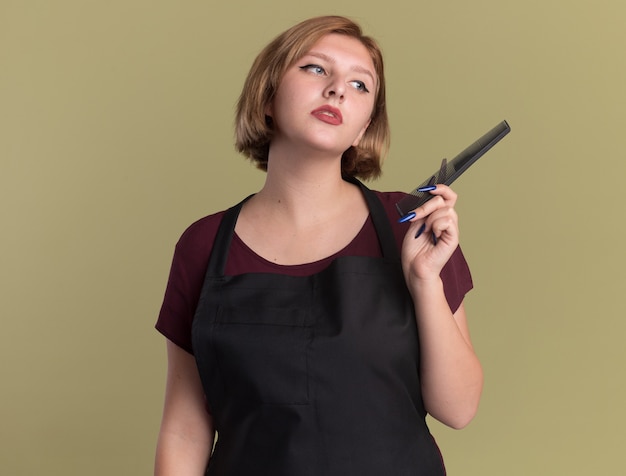 Belle jeune femme coiffeur en tablier avec des ciseaux et un peigne à cheveux à côté perplexe debout sur le mur vert