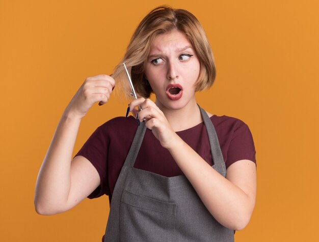 Belle jeune femme coiffeur en tablier essayant de couper ses cheveux avec des ciseaux à la confusion et surpris debout sur le mur orange