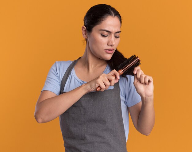 Belle jeune femme coiffeur en tablier se brosser la queue avec un visage sérieux debout sur un mur orange