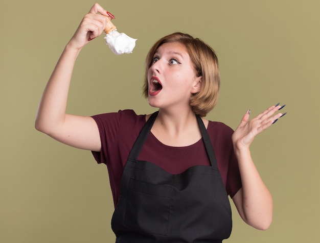 Belle jeune femme coiffeur en tablier tenant un blaireau avec de la mousse à raser en regardant étonné debout sur mur vert