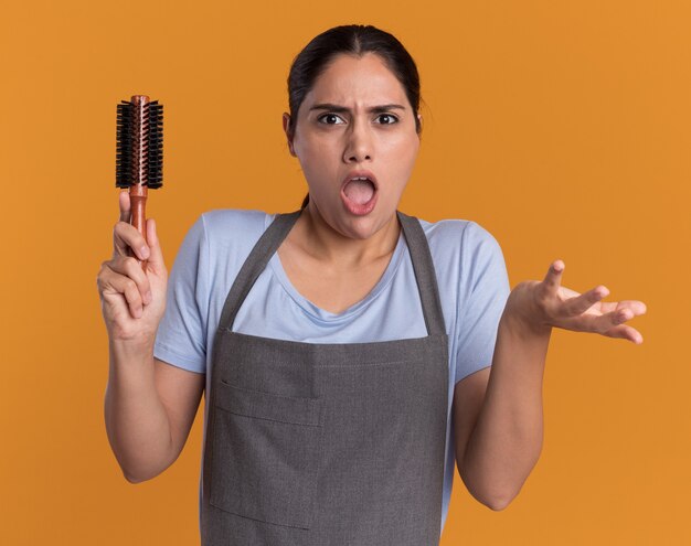 Belle jeune femme coiffeur en tablier tenant une brosse à cheveux regardant devant le bras levant confus dans le mécontentement et l'indignation debout sur le mur orange