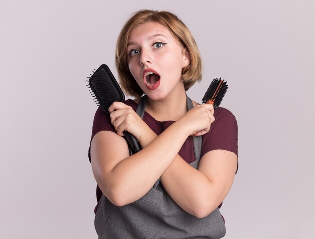 Belle jeune femme coiffeur en tablier tenant des brosses à cheveux croisant les mains à la surprise debout sur un mur blanc