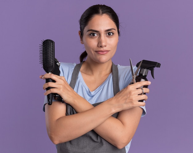 Belle jeune femme coiffeur en tablier tenant la machine de tondeuse vaporisateur et brosse à cheveux regardant à l'avant avec une expression confiante debout sur le mur violet