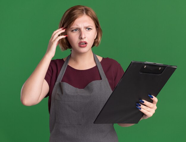 Belle jeune femme coiffeur en tablier tenant le presse-papiers à côté confus et très anxieux debout sur le mur vert