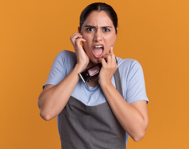 Belle jeune femme coiffeur en tablier tenant tondeuse à l'avant effrayé sur le mur orange