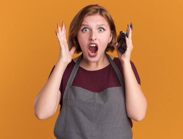 Belle jeune femme coiffeur en tablier tenant tondeuse en criant de panique avec les bras levés debout sur le mur orange