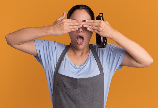 Belle jeune femme coiffeur en tablier tenant tondeuse fermant les yeux avec les mains sur le mur orange