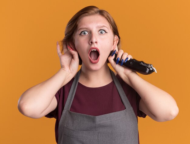 Belle jeune femme coiffeur en tablier tenant tondeuse avec les mains sur les oreilles surpris et étonné en regardant devant debout sur un mur orange
