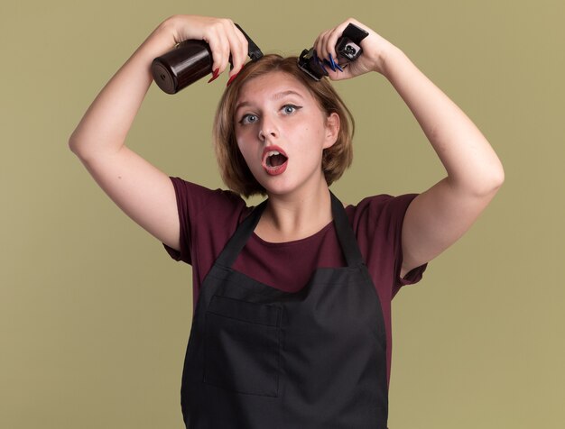 Belle jeune femme coiffeur en tablier tenant un vaporisateur et une tondeuse en essayant de couper ses cheveux debout sur un mur vert