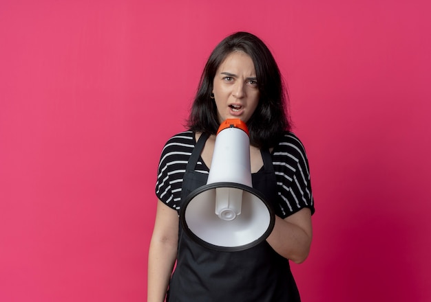 Belle jeune femme coiffeuse en tablier criant au mégaphone debout sur un mur rose