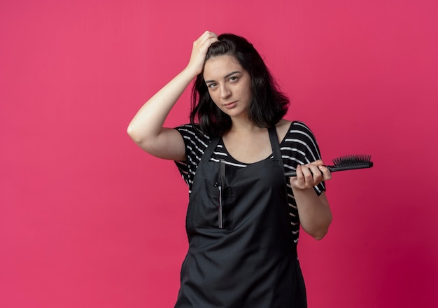 Belle jeune femme coiffeuse en tablier tenant une brosse à cheveux confus debout sur un mur rose