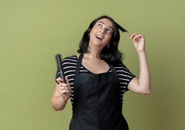 Belle jeune femme coiffeuse en tablier tenant des brosses à cheveux souriant joyeusement avec un visage heureux debout sur un mur léger