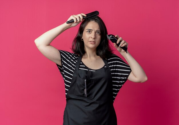 Belle jeune femme coiffeuse en tablier tenant une machine à couper les cheveux peignant ses cheveux avec une brosse debout sur un mur rose