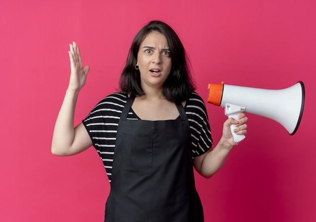 Belle jeune femme coiffeuse en tablier tenant un mégaphone confus et surpris debout sur un mur rose